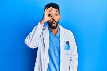 Canvas Print - Handsome hispanic man with beard wearing doctor uniform doing ok gesture shocked with surprised face, eye looking through fingers. unbelieving expression.