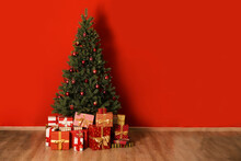 Big Beautiful Christmas Tree Decorated With Beautiful Shiny Baubles In Minimal Style And Many Different Presents On Wooden Floor. Red Wall Background With A Lot Of Copy Space For Text. Close Up.