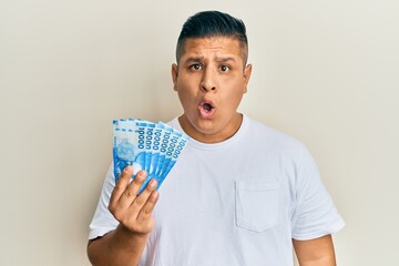 Canvas Print - Young latin man holding 10000 chilean pesos scared and amazed with open mouth for surprise, disbelief face