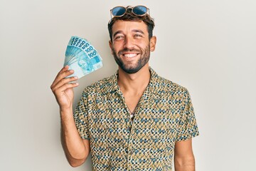 Wall Mural - Handsome man with beard holding 100 brazilian real banknotes looking positive and happy standing and smiling with a confident smile showing teeth