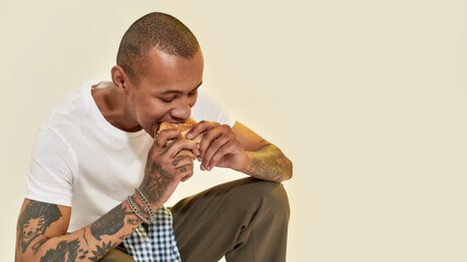 Wall Mural - Eating food. A dark-skinned tattooed man eating a bugrer sitting on his legs wearing a white t-shirt