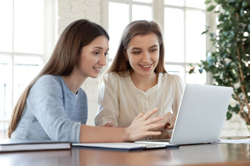 Smiling Caucasian female colleagues look at computer screen discuss company financial startup project ideas together. Happy women employees work on laptop cooperate at team meeting. Teamwork concept.