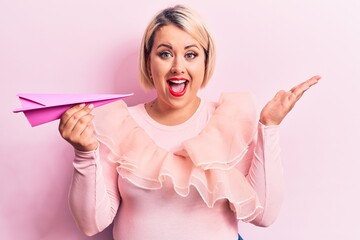 Wall Mural - Young beautiful blonde plus size woman holding paper airplane over isolated pink background celebrating achievement with happy smile and winner expression with raised hand