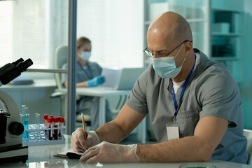Poster - Mature bald male scientist in protective mask and gloves making scientific notes