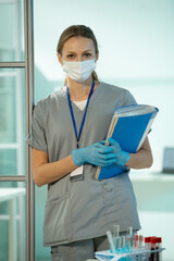 Poster - Young clinician in uniform, protective mask and gloves holding medical documents