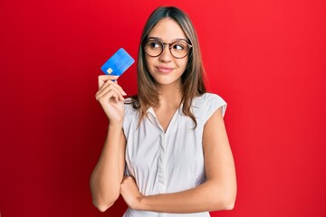 Young brunette woman holding credit card smiling looking to the side and staring away thinking.