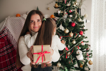 Young woman holding a present and hugging her daughter in front of the New Year tree. Families on New Year concept.