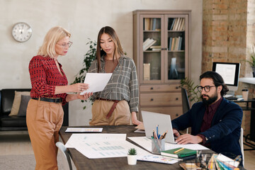 Poster - Young and mature females standing by table and discussing sketch on paper