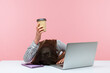 © khosrork - Sleepy bored woman office worker lying on table with laptop holding and showing paper coffee cup, feeling lack of energy sitting at workplace. Indoor studio shot isolated on pink background