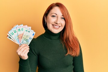 Wall Mural - Beautiful redhead woman holding euro banknotes looking positive and happy standing and smiling with a confident smile showing teeth