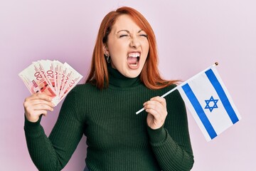 Sticker - Beautiful redhead woman holding 20 shekels banknotes and israel flag angry and mad screaming frustrated and furious, shouting with anger. rage and aggressive concept.