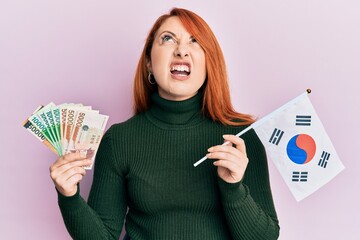 Poster - Beautiful redhead woman holding south korean won banknotes and flag angry and mad screaming frustrated and furious, shouting with anger looking up.