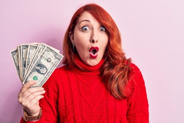 Canvas Print - Young beautiful redhead woman holding bunch of dollars banknotes over pink background scared and amazed with open mouth for surprise, disbelief face