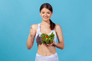 Wall Mural - Healthy positive woman in white sportswear with perfect slim body showing thumbs up holding big bowl with fresh vegetable salad, keeping detox diet. Indoor studio shot isolated on blue background