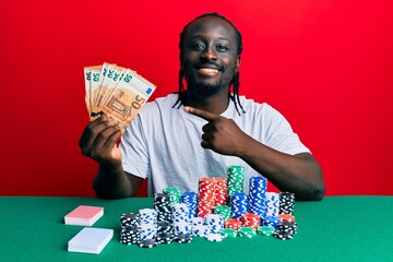 Poster - Handsome young black man playing poker holding 50 euros banknotes smiling happy pointing with hand and finger