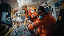 Female And Male EMS Paramedics Provide Medical Help To An Injured Patient On The Way To A Healthcare Hospital. Emergency Care Assistants Putting On Non-Invasive Ventilation Mask In An Ambulance.