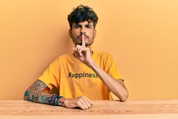 Poster - Young hispanic man wearing tshirt with happiness word message sitting on the table asking to be quiet with finger on lips. silence and secret concept.