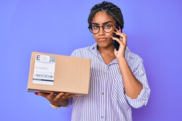 Wall Mural - Young african american woman with braids holding delivery box calling assistance clueless and confused expression. doubt concept.