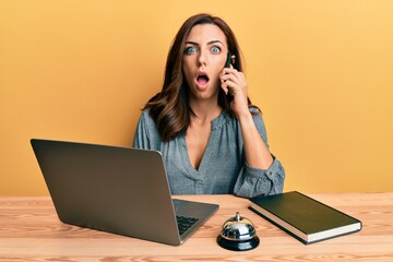 Wall Mural - Young brunette woman working at hotel reception talking on the phone afraid and shocked with surprise and amazed expression, fear and excited face.