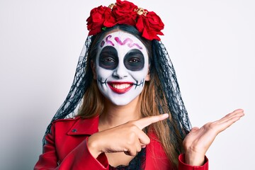 Wall Mural - Woman wearing day of the dead costume over white amazed and smiling to the camera while presenting with hand and pointing with finger.