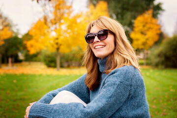 Poster - Portrait shot of happy mature woman wearing sunglasses and turtleneck sweater while relaxing in the park at autumn.