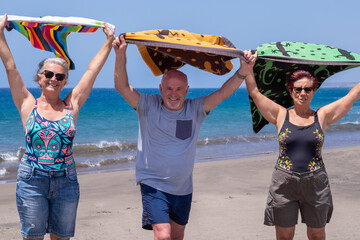 Three senior friends enjoying beach together holding towels in the wind, smiling with blue sea in background - active retired seniors and vacation concept