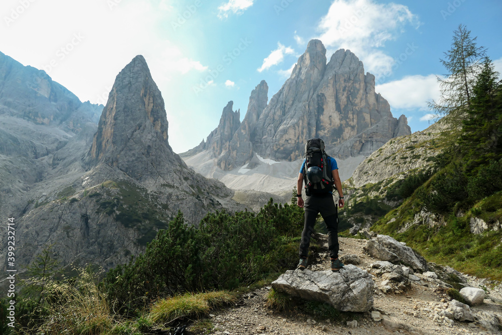 A man with big backpack hiking in high, Italian Dolomites. There are many sharp peaks behind. He is standing on a big boulder, enjoying the view. There are a few trees around. Sunny day. Outdoor - obrazy, fototapety, plakaty 