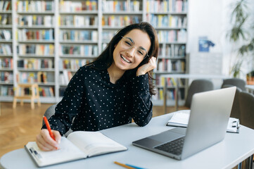 Wall Mural - Portrait of an amazing caucasian young smiling woman, is sitting at the desk while remote work from home using laptop and looks into a camera