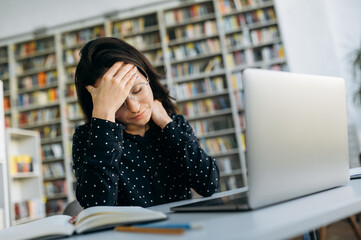 Wall Mural - Headache while working. Stress. Young caucasian woman, manager or student, upset and tired from work sits at the desk, holding her head