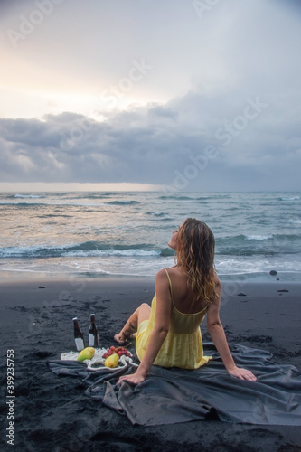 A beautiful blonde girl in a yellow dress sits on the beach with black sand and looking at sunset. Romantic setting for a fruit picnic by the ocean © Serge Touch