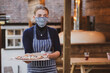 © Mint Images - Woman waitress in apron and face mask holding plate of pizza.