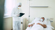 © My Ocean studio - An adult male patient lies on a bed in a hospital ward while he is examined by a doctor in protective clothing and a mask.