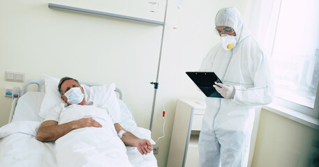 Wall Mural - An adult male patient lies on a bed in a hospital ward while he is examined by a doctor in protective clothing and a mask.