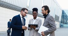 African American Young Businesswoman And Two Caucasian Businessmen Standing At Street And Discussing Work With Tablet Device In Hands. Mixed-races Woman And Men Talking About Working Project.