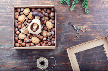 Carton box with two kind of nuts in shell: walnuts, hazelnuts and mushroom shaped nutcracker, scissors, jute thread, on rustic wooden table. Healthy Christmas gift concept.