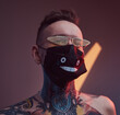 © Fxquadro - Dark and atmospheric room with colourful walls and spotlights and cheerful tattooed guy with naked torso and modern hairstyle weared with mask and sunglasses.