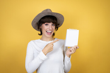 Young caucasian woman wearing hat over isolated yellow background smiling, surprised and pointing blank notebook in her hand