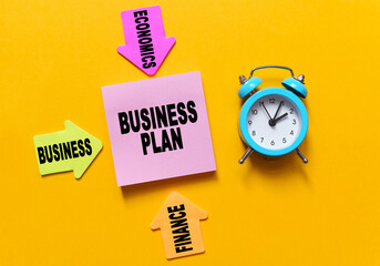 On a yellow background, a blue alarm clock, multi-colored paper arrows with inscriptions and paper for notes on which the text is written - BUSINESS PLAN