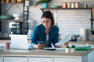 Concentrated young business woman using her mobile phone while working with computer in the kitchen at home.