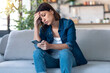 © nenetus - Worried young woman using her mobile phone while thinking about problems sitting on couch in the living room at home.