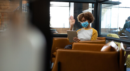 Wall Mural - Mixed race woman wearing face mask using tablet and earphones waving during video meeting