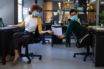 Wall Mural - Mixed race woman and man wearing face masks passing document in casual office