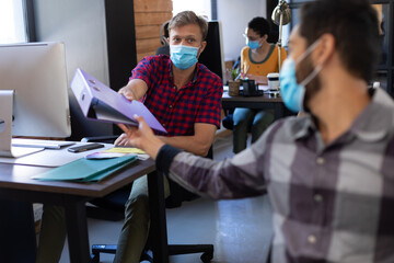 Wall Mural - Diverse male colleagues wearing face masks passing file between desks