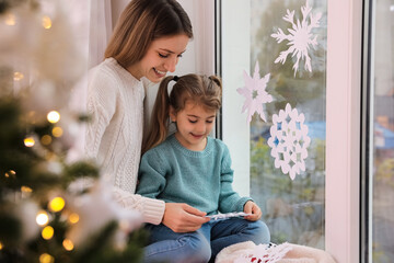 Wall Mural - Happy mother and daughter near window decorated with paper snowflakes indoors