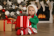 Cute Baby In Santa's Elf Clothes Holding Gift Box Near Christmas Tree At Home