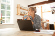 © PETE  MULLER/Westend61 - Senior woman looking away while drinking coffee sitting with laptop in kitchen at home