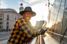 Smiling African Man With Smart Phone Looking Away While Standing In City During Sunset