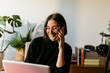 © VALENTINA BARRETO STUDIO/Westend61 - Smiling businesswoman talking on smart phone while using laptop sitting at home