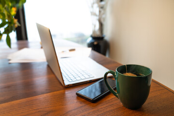 Work from home remote work station background image with laptop set up on a wood dining table with cell phone, papers and hot tea and light coming in from a window beyond.