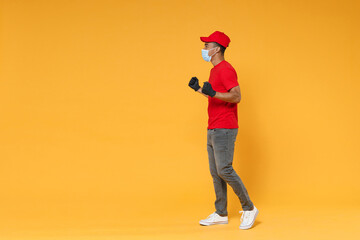 Wall Mural - Full length delivery employee african man in red cap t-shirt face mask gloves uniform work courier dealer service on quarantine coronavirus covid-19 virus concept isolated on yellow background studio.